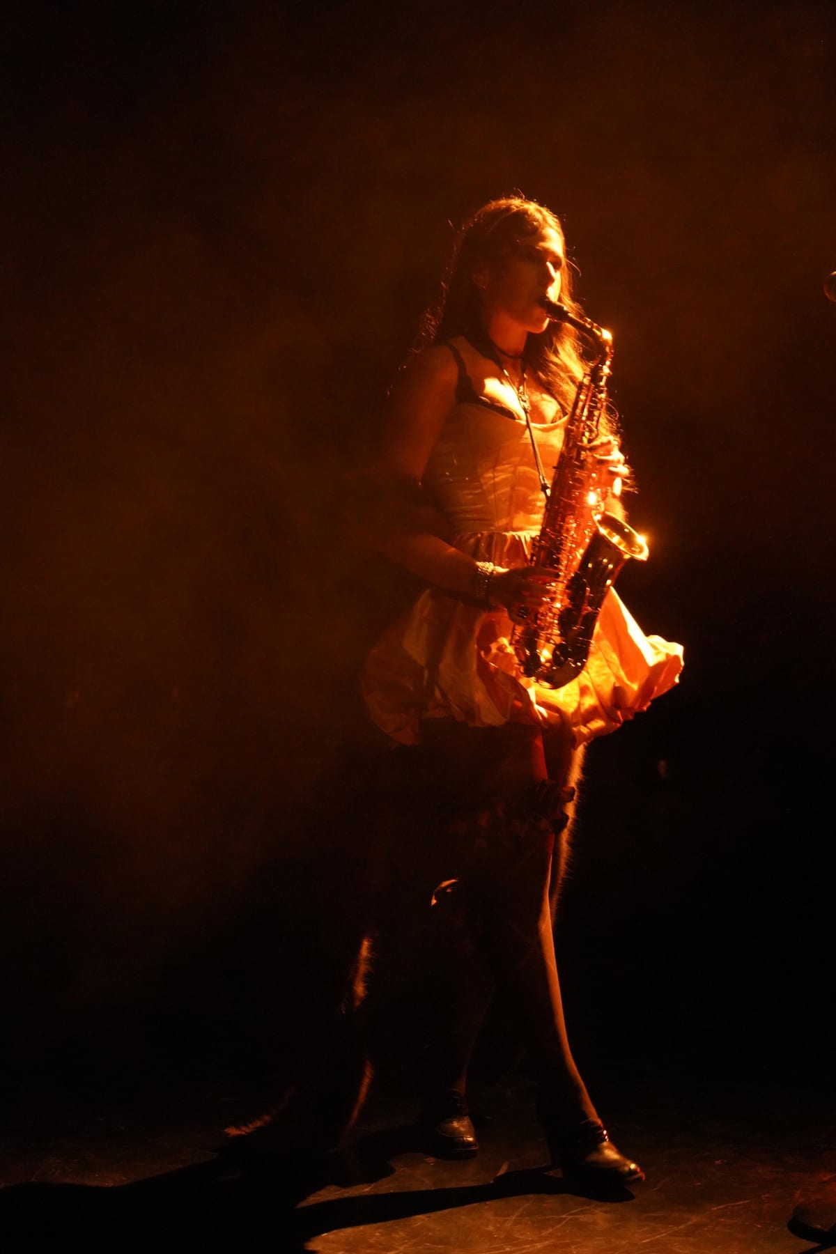 Saxophonistin im warmen Bühnenlicht, das Instrument schräg im Anschnitt. Konzentration auf dem Spiel, dunkler Hintergrund.